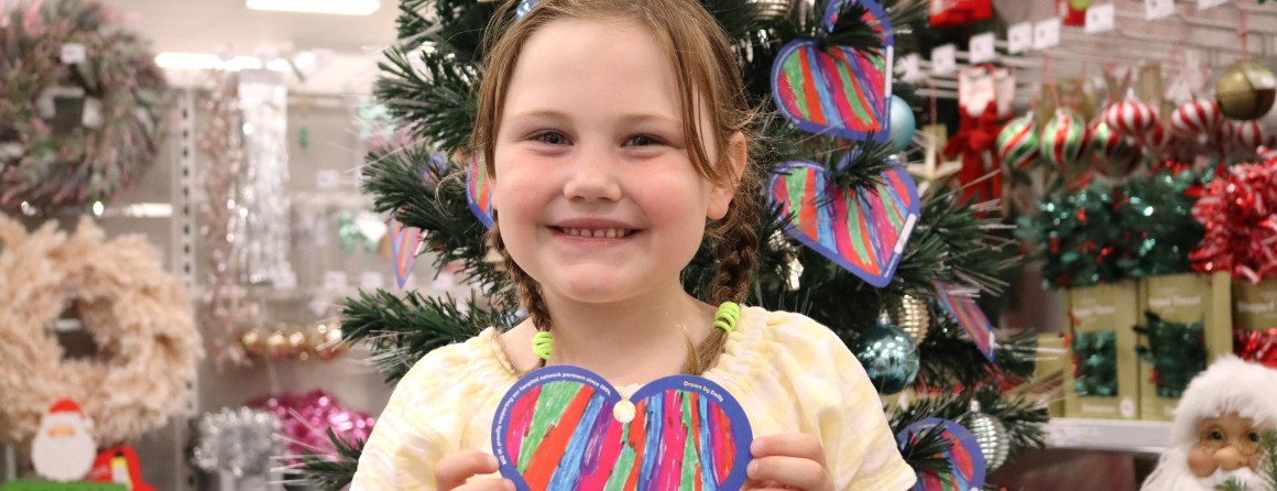 A young girl with light brown hair grins at the camera, holding a colourful heart that she designed. She wears a yellow dress, has her hair in plaits and stands in front of a Christmas tree.